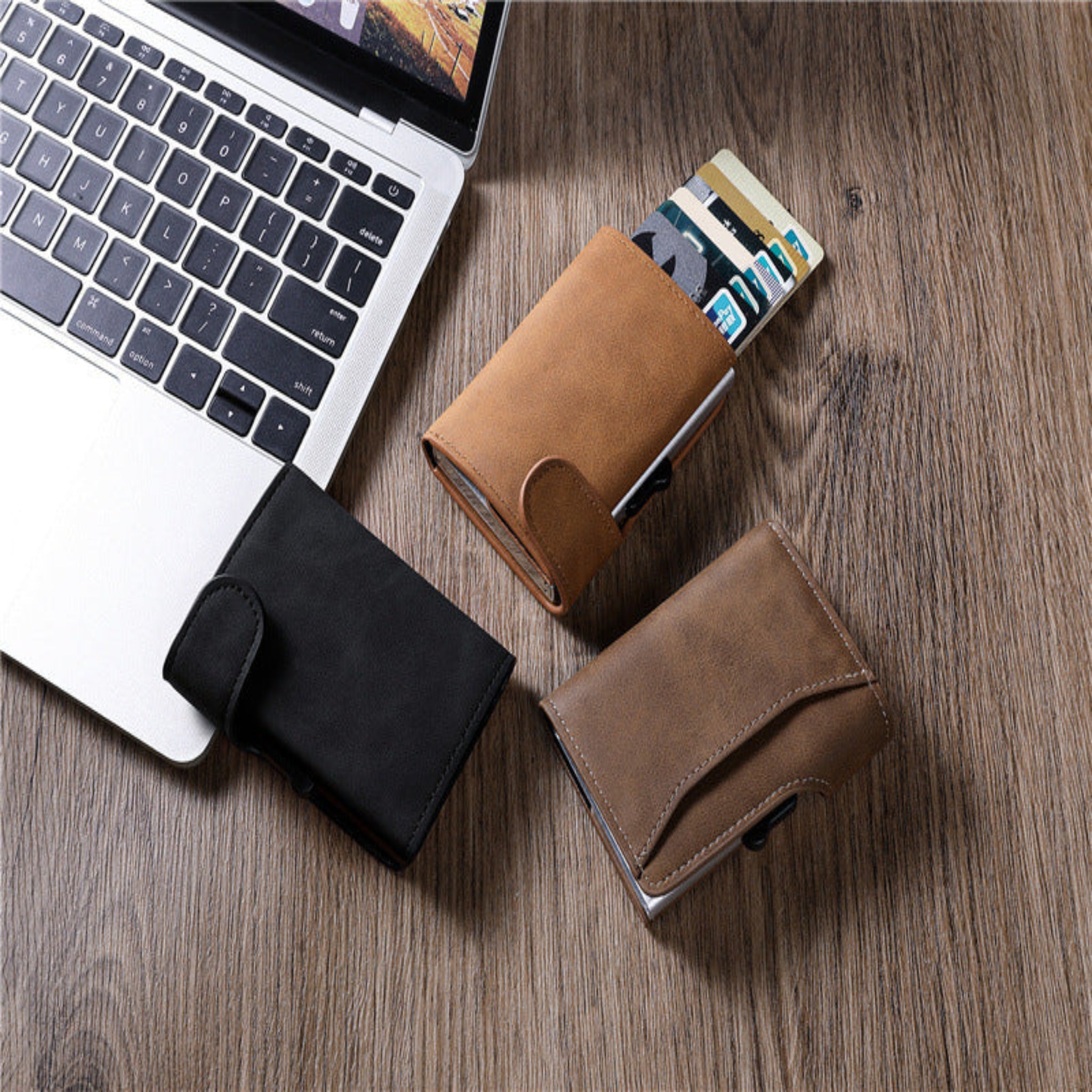 Three leather wallets in black, brown, and tan on a wooden surface with a laptop.
