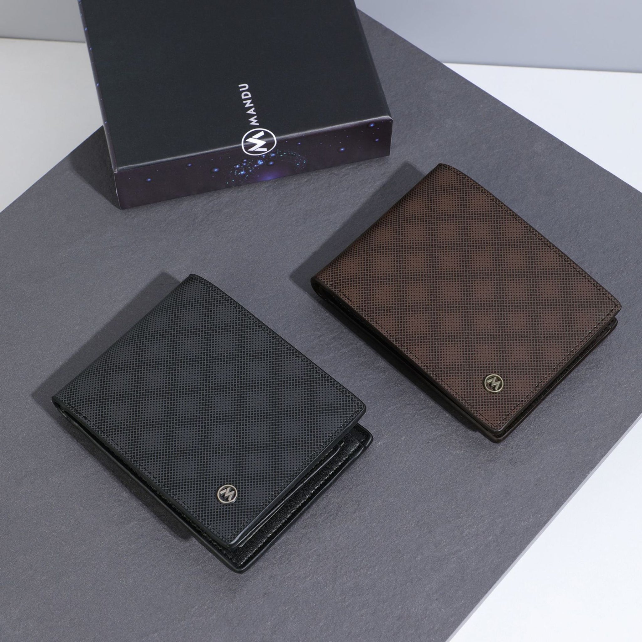 Two wallets, one black and one brown, on a gray surface with a brand box in the background.